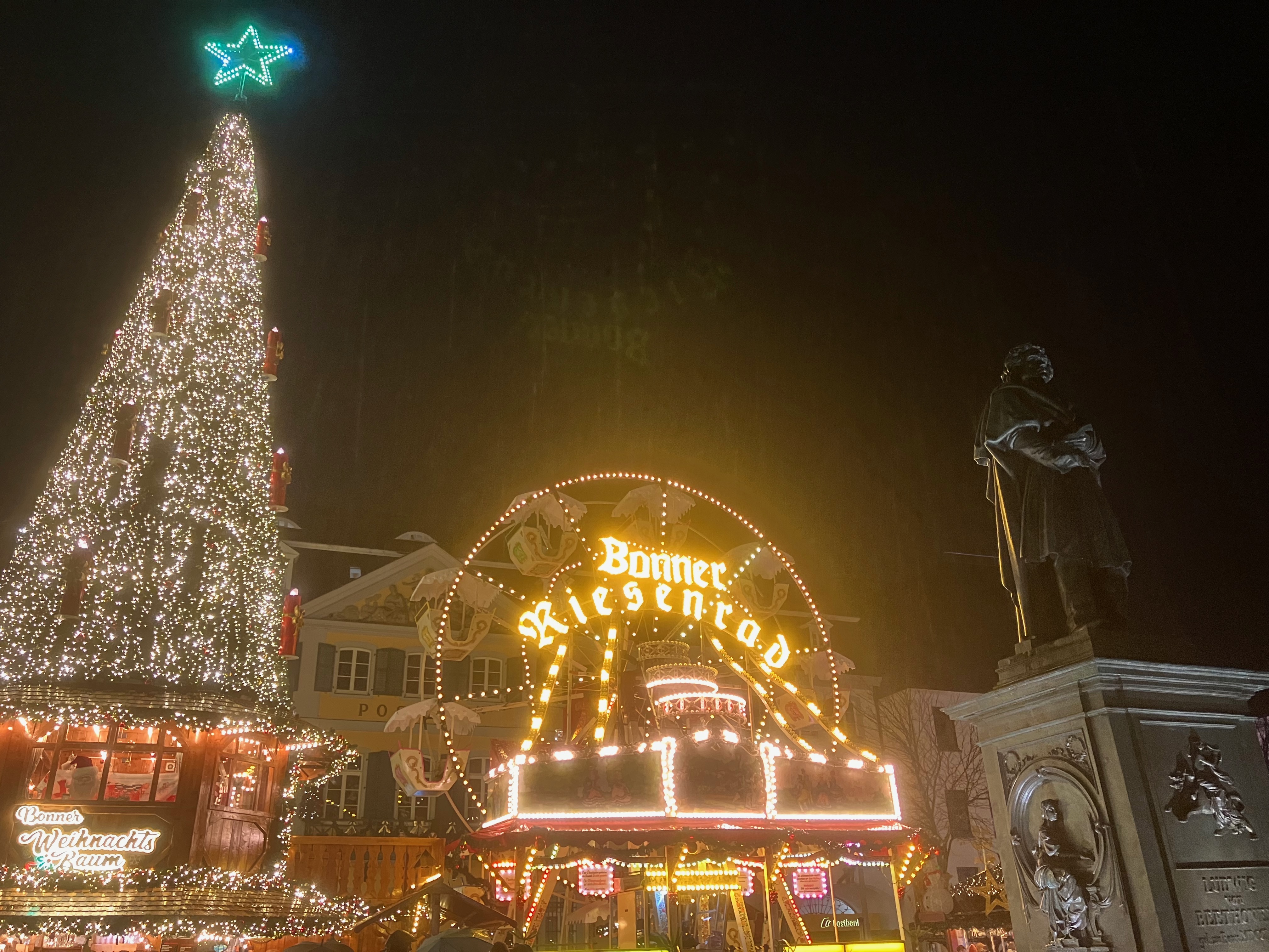 A statue of Beethoven is watched over by the Bonn Christmas Market wheel and tree in Bonn. It is nighttime and the wheel and tree are very brightly lit.
